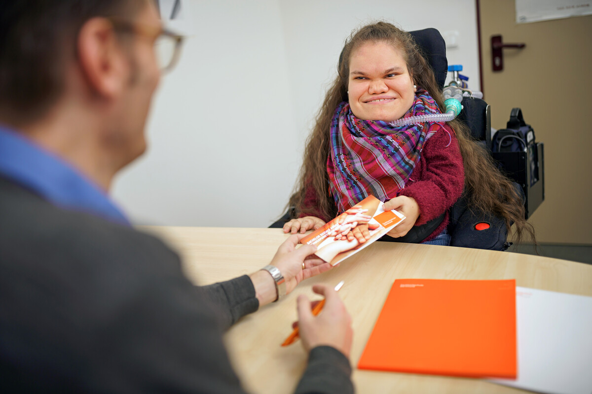 Beratungsgespräch zu barrierefreiem Studieren an der FH Dortmund: Ein Berater übergibt eine Broschüre an eine Studentin im Rollstuhl.