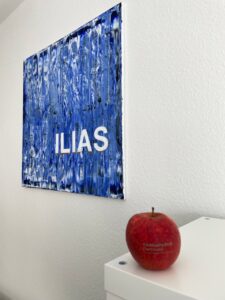 Foto: Im Vordergrund ein Apfel mit der Aufschrift Fachhochschule Dortmund, links im Hintergrund ein Gemälde des blauen ILIAS Logos