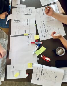 Foto von einer Kleingruppen-Arbeit, auf dem man den Tisch voller Papiere, Post-Its, Stifte und Kleber sieht.