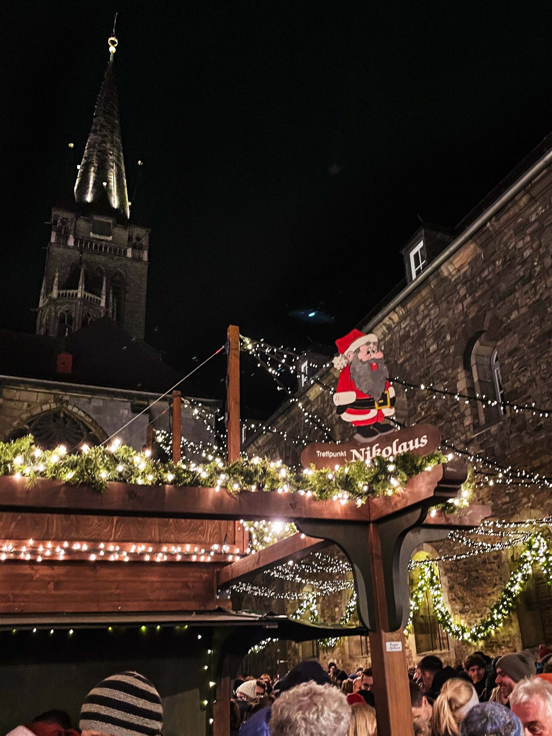 Weihnachtsmarkt Aachen. Personen und Weihnachtsstände sind zu erkennen. Ein Schild mit Treffpunkt Nikolaus ist dominiert das Bild.