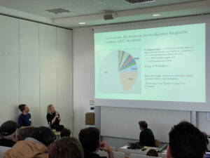 Fabian Kruse und Kendra Grotz beim Vortrag, rechts Leinwand mit Präsentation mit dem Text "Generisches Maskulinum durch inklusive Ansprache"