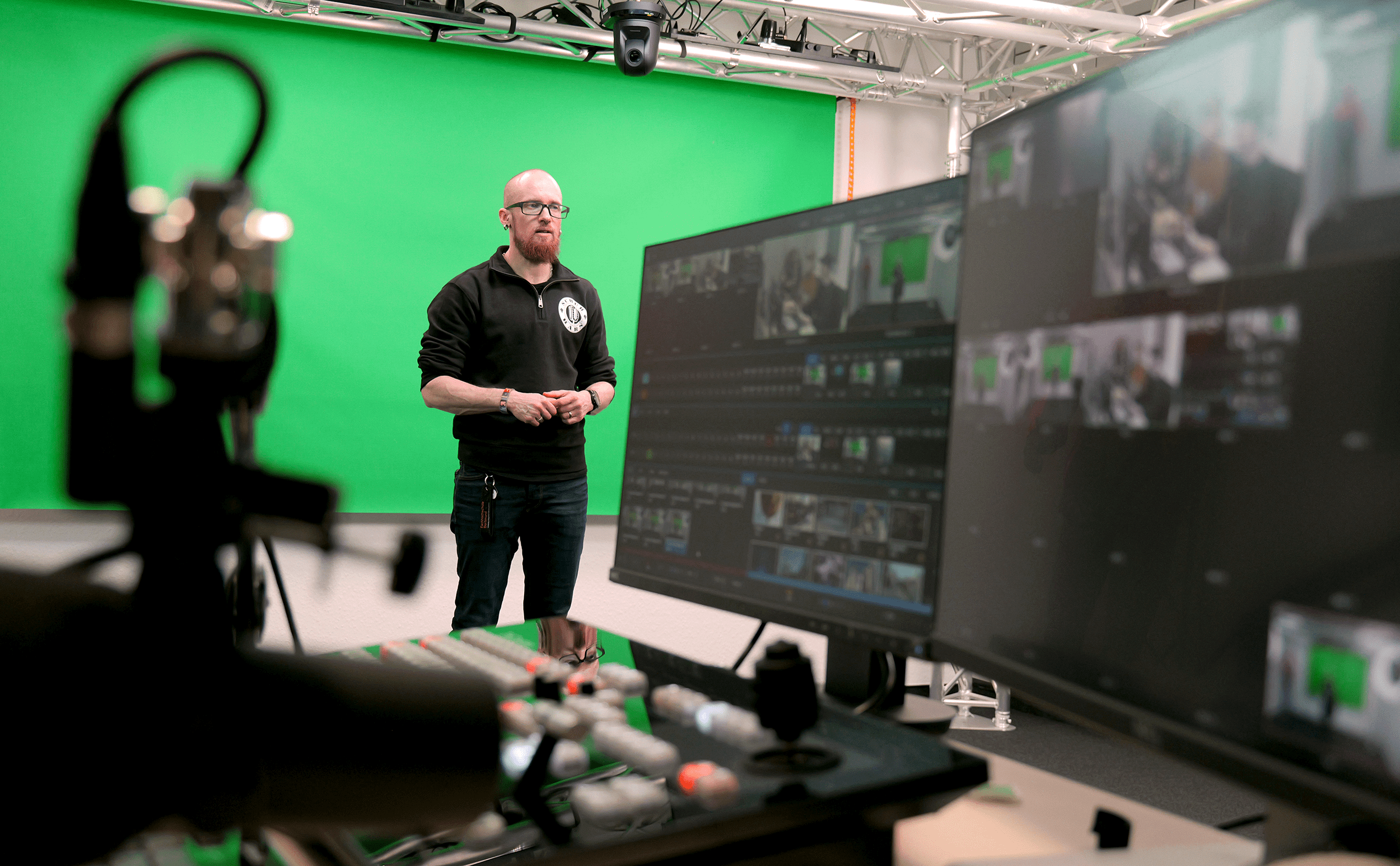 Vincent Beringhoff steht im Studioraum des Videolabors vor einem Greenscreen, im Vordergrund ein Regiepult mit zwei Monitoren