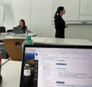 Aufgeklappter Laptop, gezeigt wird das Etherpad in ILIAS, im Hintergrund Kseniia Gribchenko und Elyesa Seidel beim Vortrag