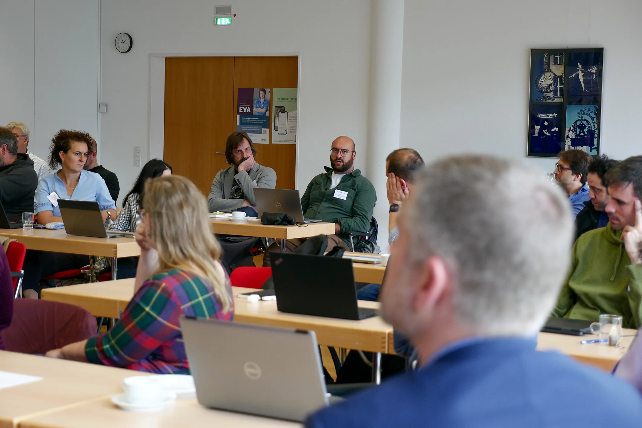 Unscharf im Vordergrund: Hinterkopf eines Teilnehmenden. Im Hintergrund: Teilnehmende an Tischen sitzend, ein Teilnehmer spricht, die anderen hören zu.