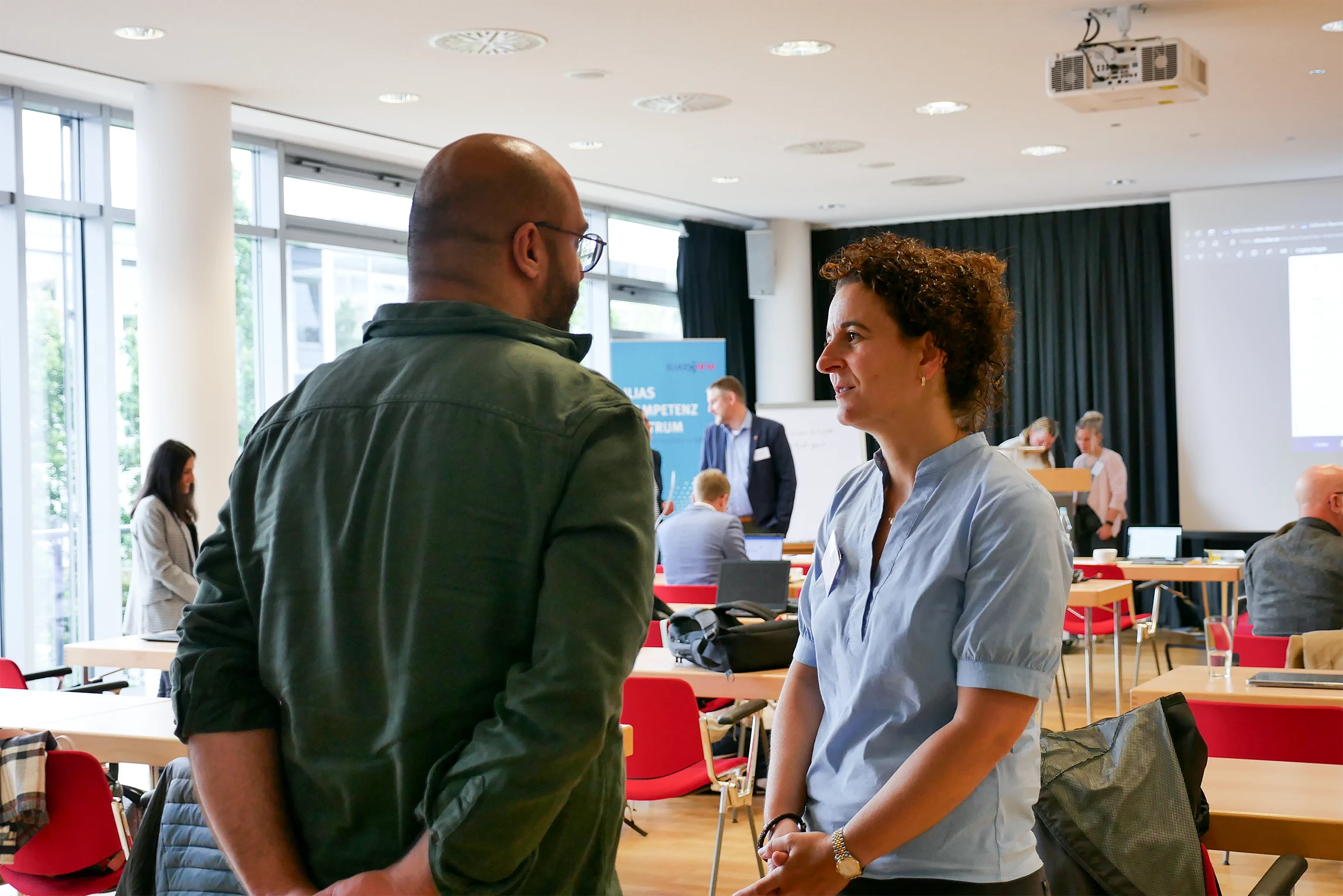 Im Vordergrund: Zwei Teilnehmende stehend, im Gespräch. Im Hintergrund Vortragsraum mit weiteren Teilnehmenden.