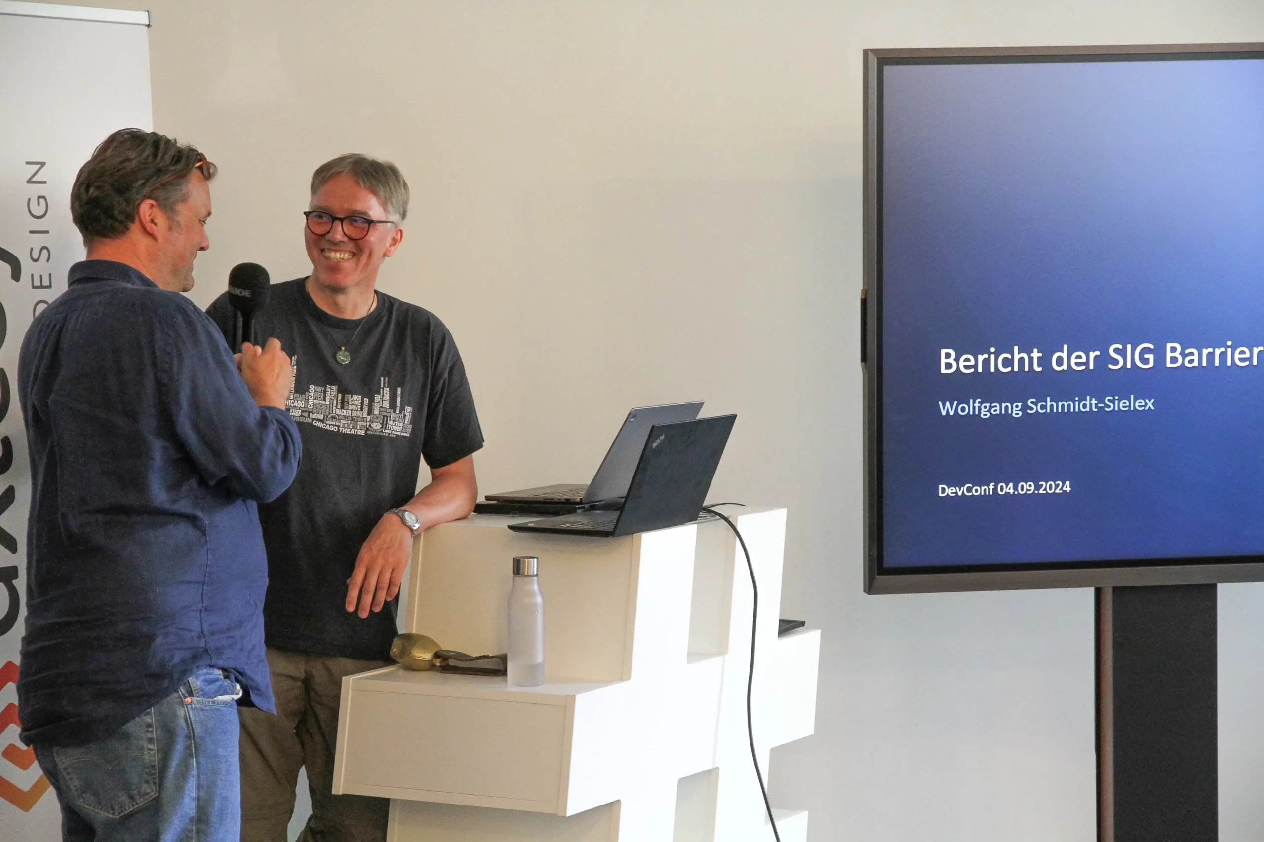 Foto: Zwei Vortragende am Rednerpult unterhalten sich, rechts neben ihnen ein Monitor mit der Präsentation, nur teilweise ist der Text zu sehen: "Bericht der SIG Barrier[efreiheit], Wolfgang Schmidt-Sielex"