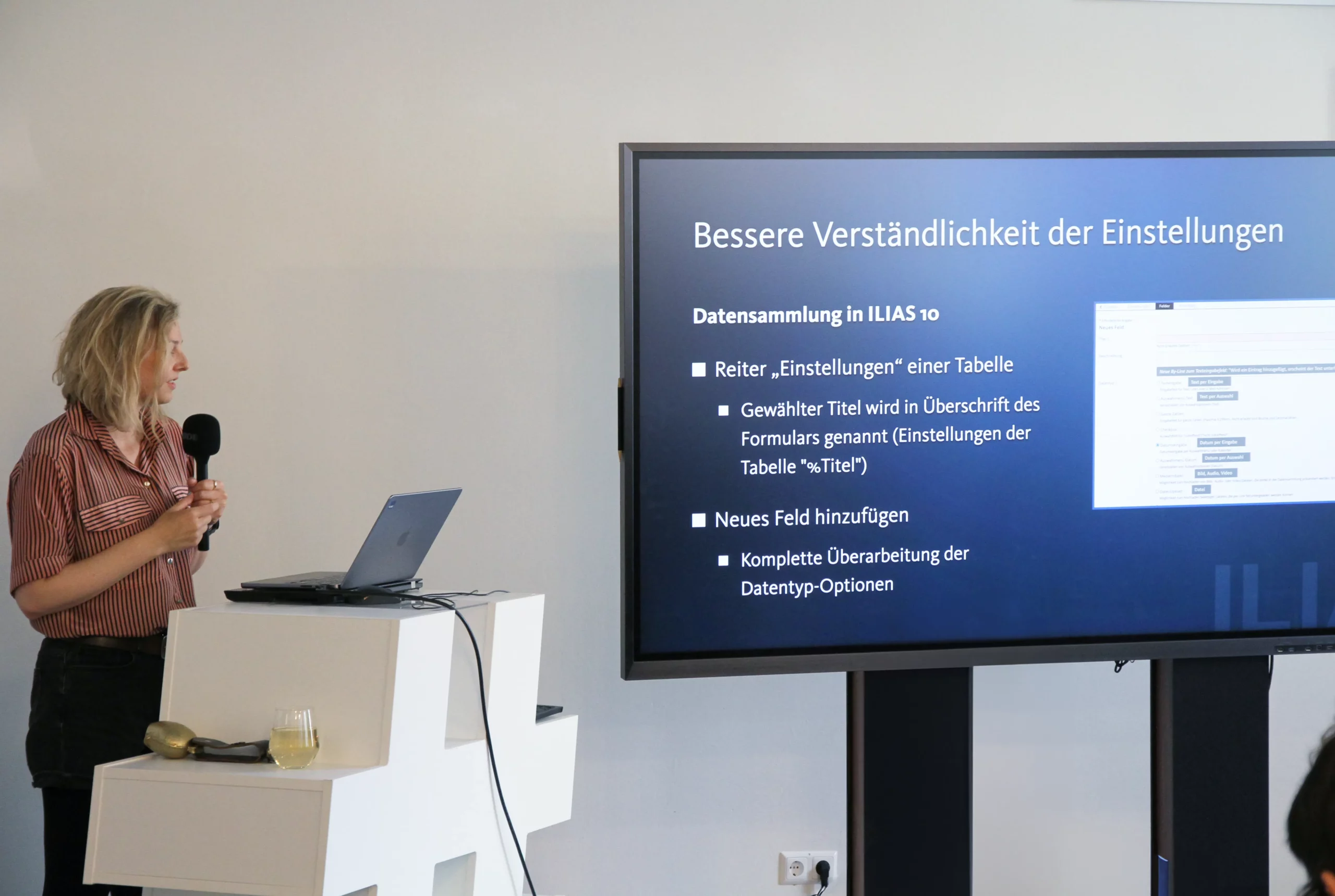 Foto: Kendra Grotz beim Vortrag, rechts auf einem Monitor ihre Präsentation mit dem Text "Bessere Verständlichkeit der Einstellungen"