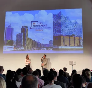 Foto: Vortragssituation, zwei Personen auf der Bühne, Text auf der präsentierten Folie: "25. ILIAS Konferenz 10. & 11. September 2026 Hamburg".