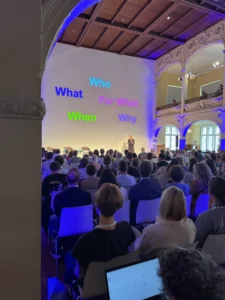 Foto: Vortragssituation, im Vordergrund ist das Publikum von hinten zu sehen. Hinter der Vortragenden sind an der Wand die Worte "Who", "What, "For What", "Why" und "When" zu lesen.