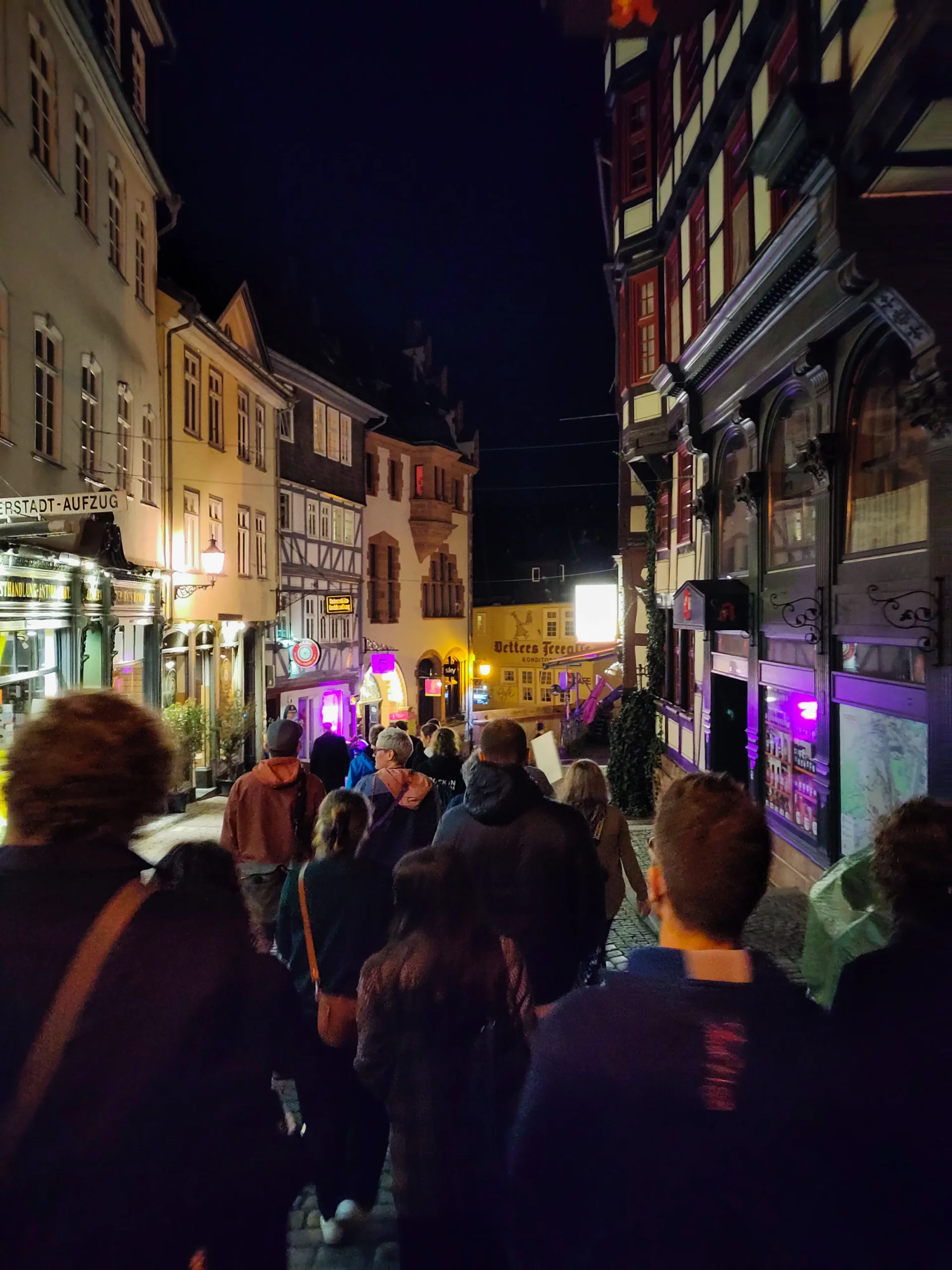 Foto: Eine Gruppe von Personen geht nachts eine Straße mit historischen Gebäuden und beleuchteten Schildern entlang.