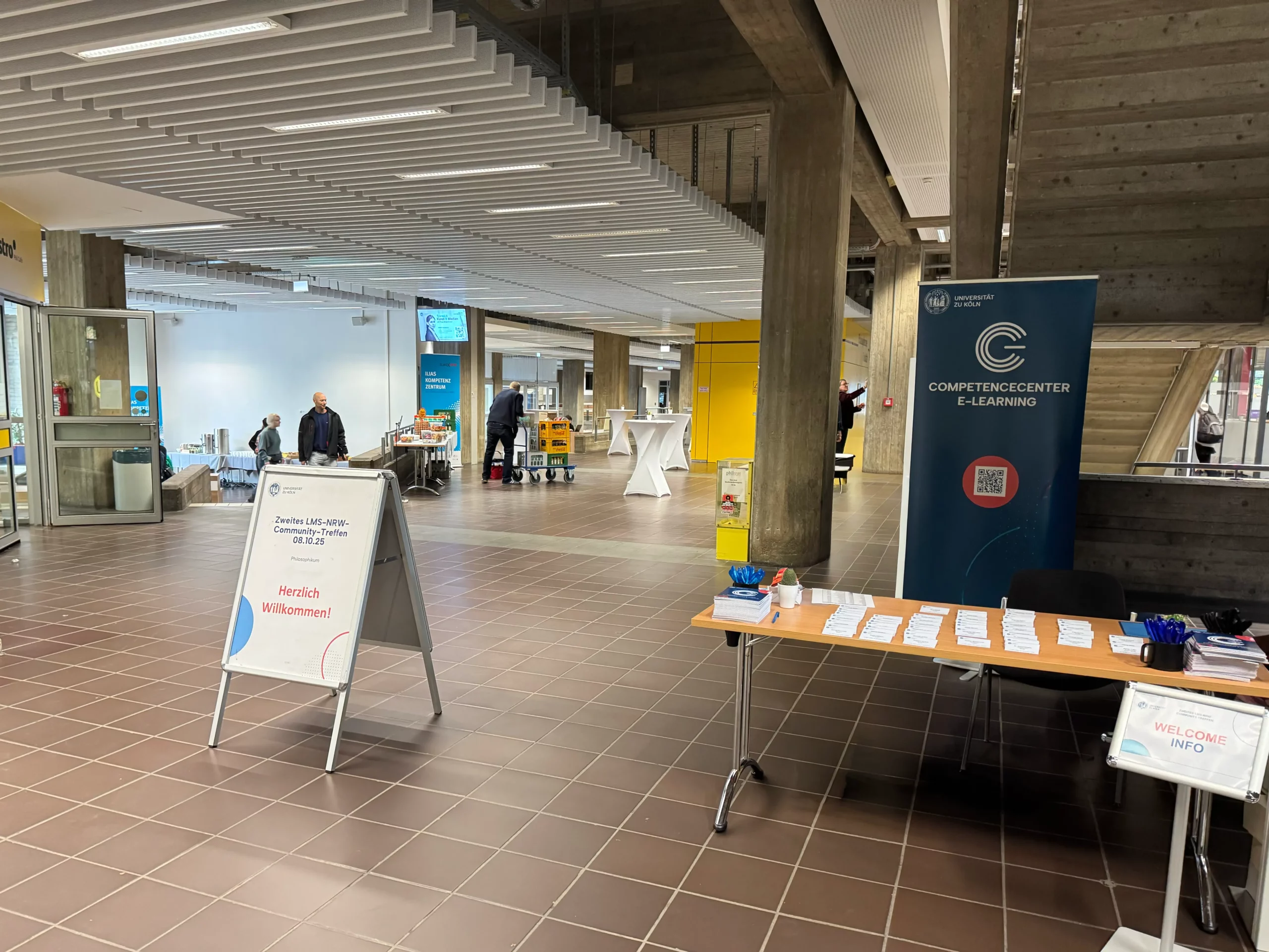 LMS Köln_2 Foto: Innenraum mit Informationsstand und Stehtischen im Hintergrund, auf einem Schild steht "Herzlich Willkommen!" und "Zweites LMS-NRW-Community-Treffen" , auf einem Banner "Competence Center E-Learning".