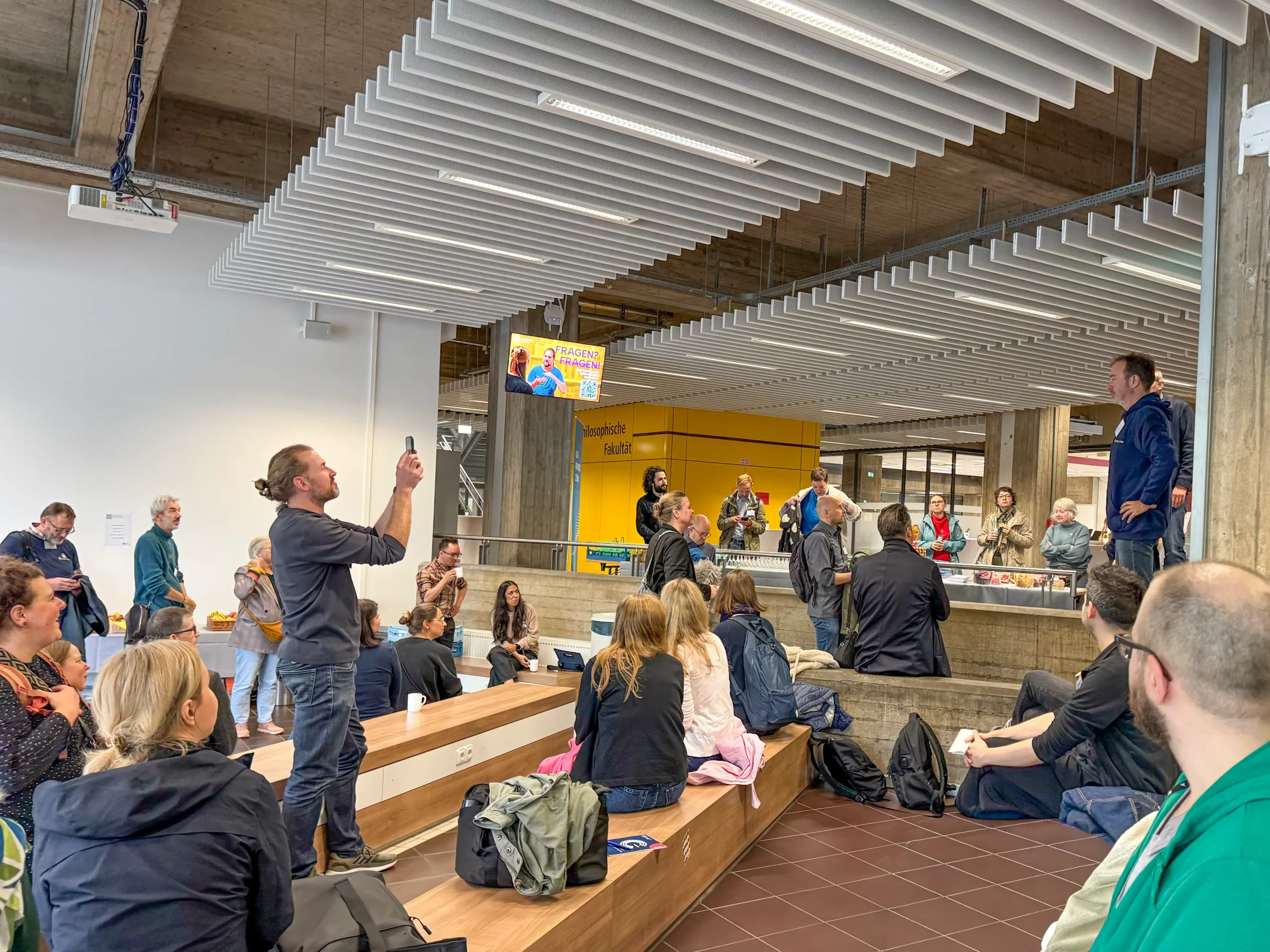 LMS Köln_3 Foto: Personen sitzen und stehen in einem Foyer mit Holzsitzbänken und sichtbaren Betonsäulen und hören einem stehenden Mann zu. Ein anderer Mann hält stehend sein Handy hoch.