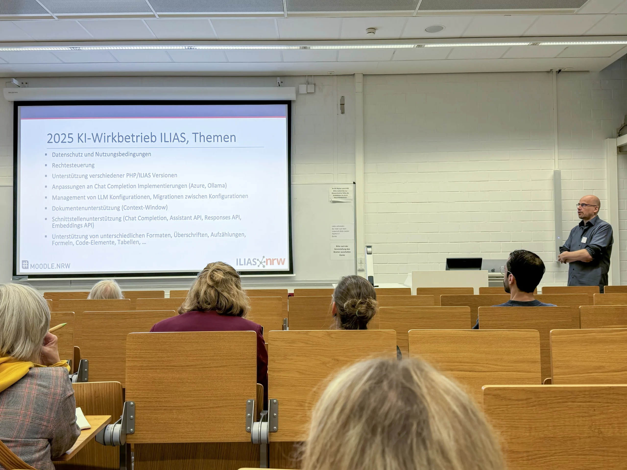 LMS Köln_4 Foto: Vortragender steht neben einer Leinwand mit Präsentationsfolie zu KI-Wirkbetrieb ILIAS 2025, Publikum sitzt in Hörsaal mit Holzsitzen.