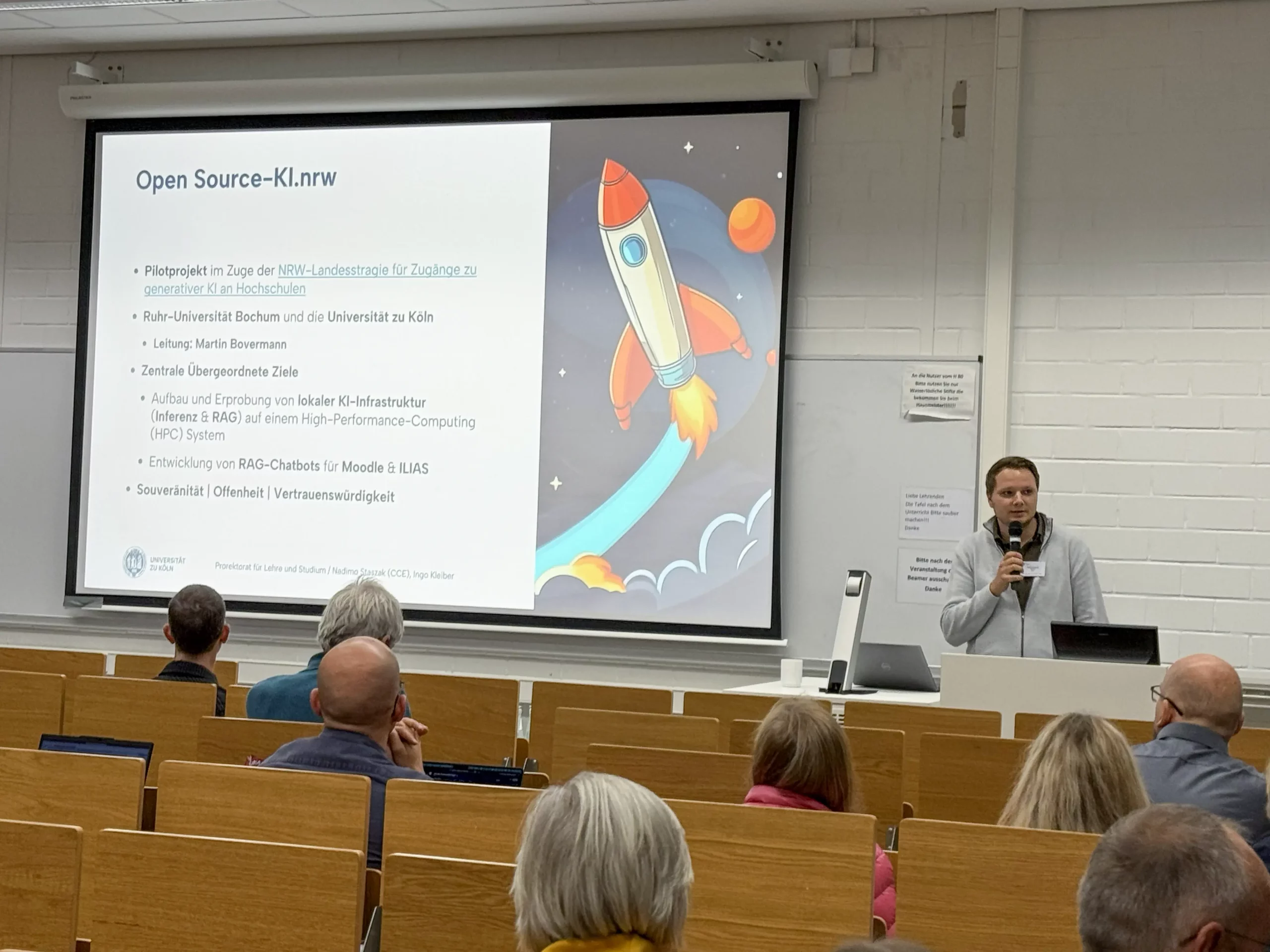 LMS Köln_6 Foto: Vortragender mit Mikrofon vor Leinwand mit Präsentation zu Open Source-KI.nrw, die eine bunte Rakete im Start zeigt; Publikum im Hörsaal.