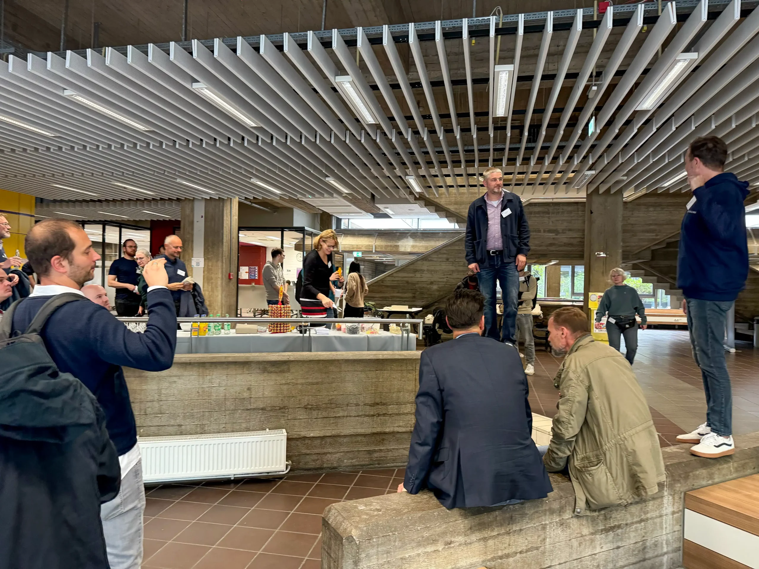 LMS Köln_9 Foto: Mehrere Personen in einem Foyer, einige sitzen auf einer kleinen Mauer, andere stehen und unterhalten sich.