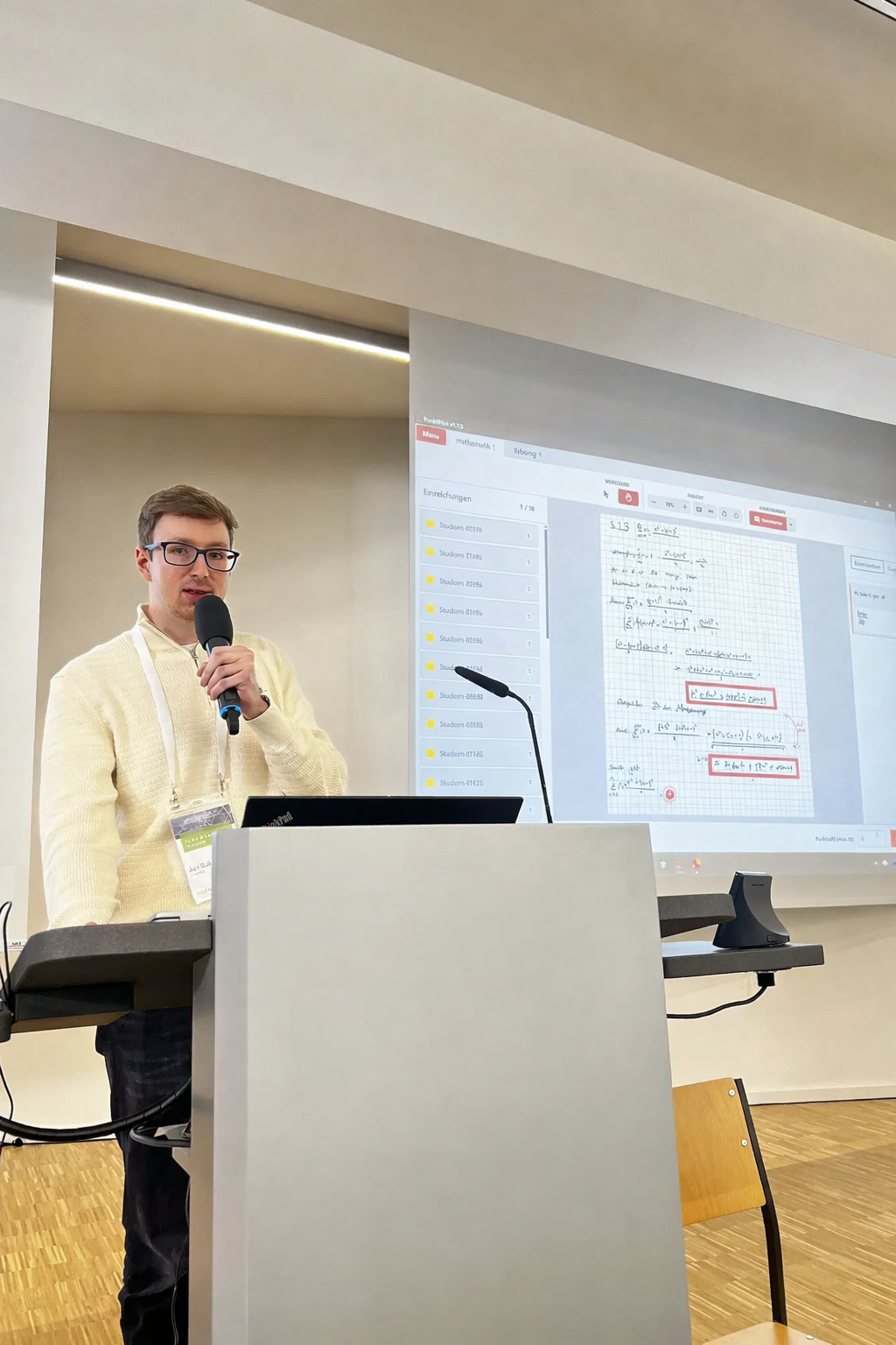 Foto: Ein Redner mit einem Mikrofon in der Hand an einem Rednerpult. Neben ihm eine Leinwand mit einer Präsentation.