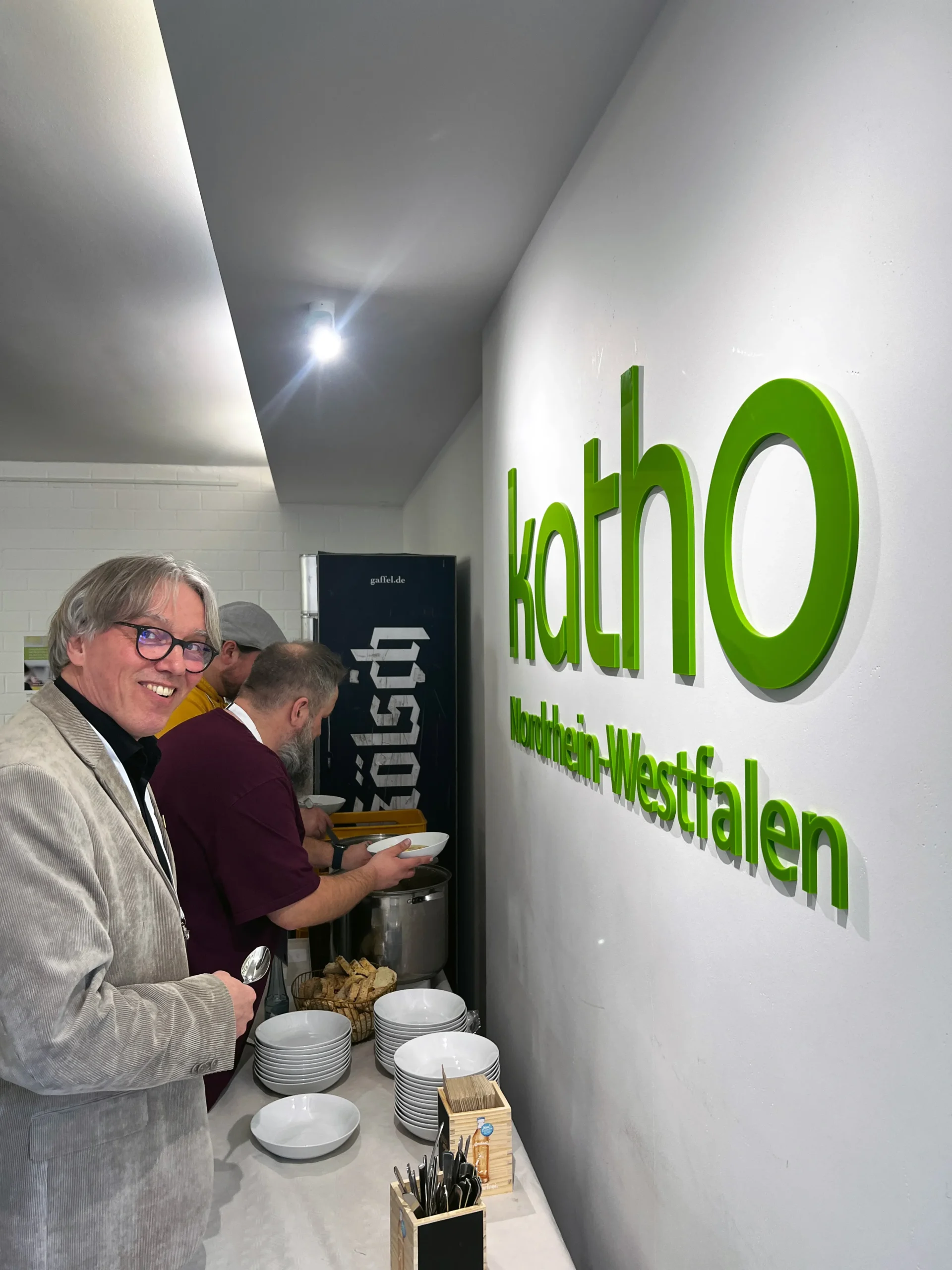 Foto: Drei Personen stehen an einem Buffet mit Tellern und Besteck vor einer Wand mit dem grünen Schriftzug »katho Nordrhein-Westfalen«