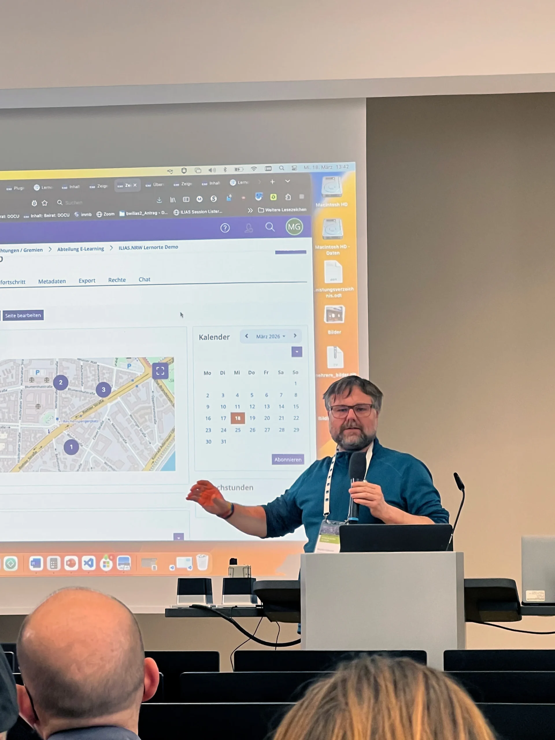 Foto: Ein Redner mit einem Mikrofon in der Hand an einem Rednerpult. Hinter ihm eine Leinwand mit einer Präsentation.