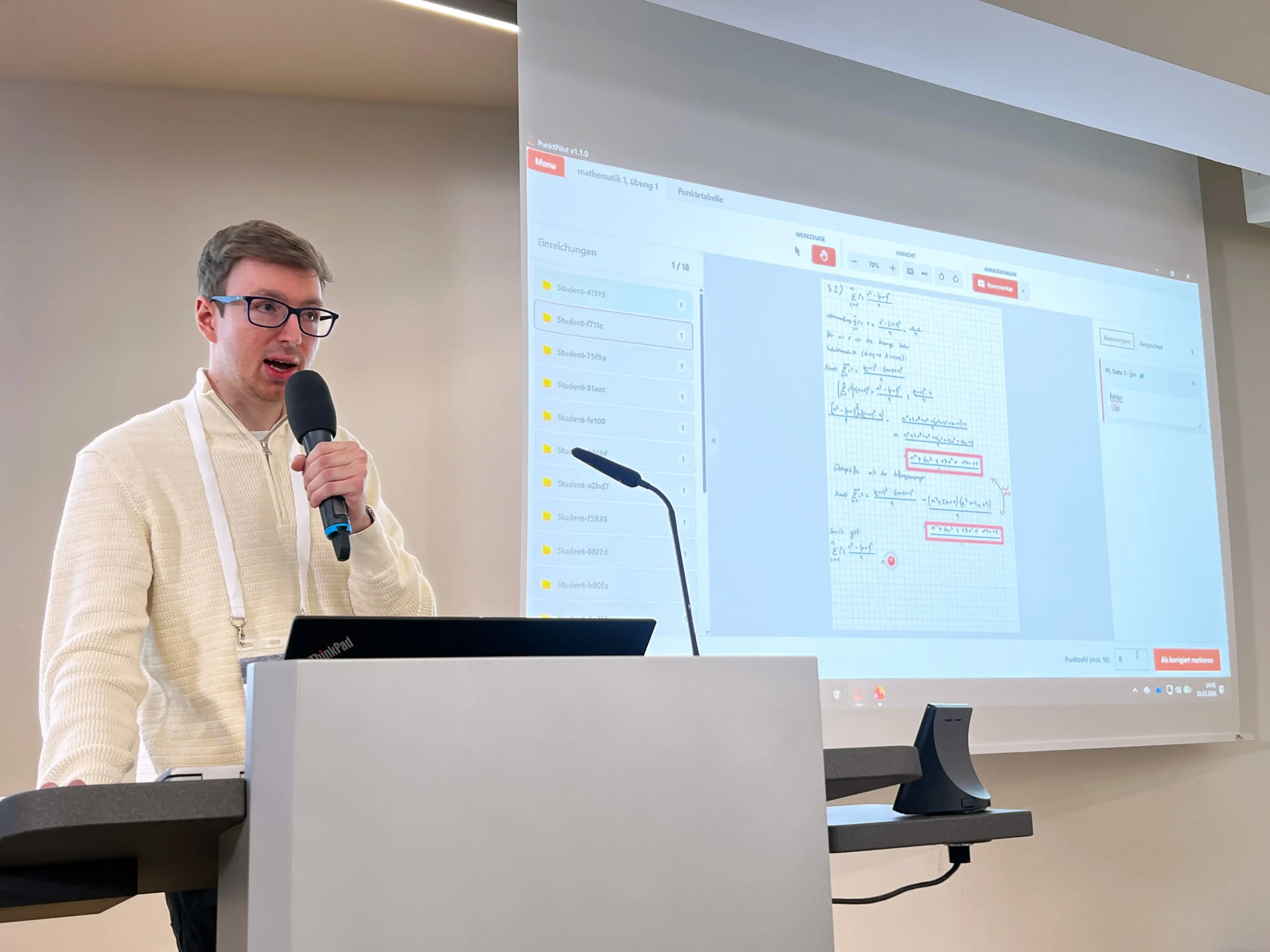 Foto: Ein Redner mit einem Mikrofon in der Hand an einem Rednerpult. Neben ihm eine Leinwand mit einer Präsentation.