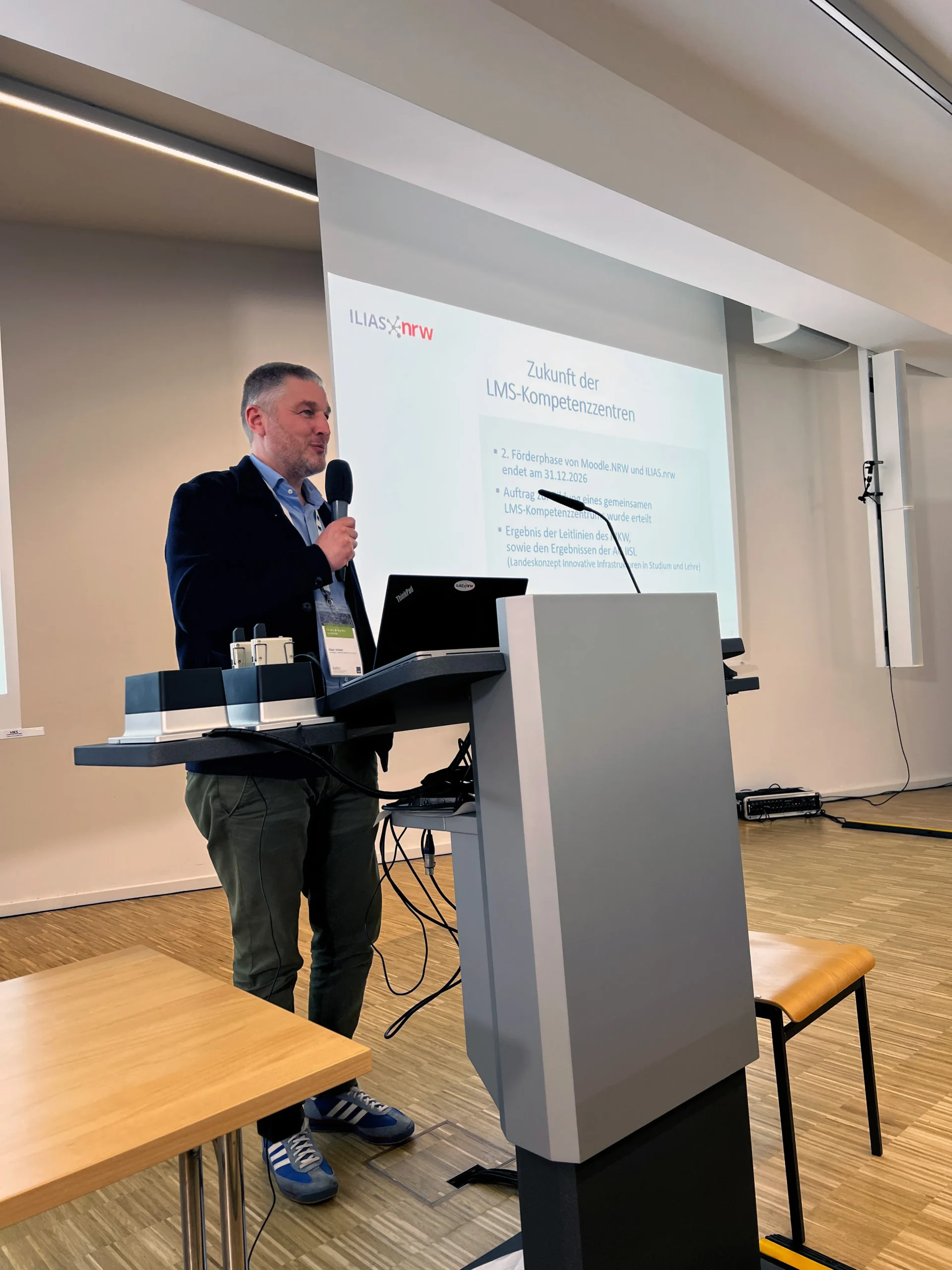 Foto: Vortragender an einem Rednerpult mit einem Mikro in der Hand, hinter ihm eine Leinwand mit dem Text "Zukunft der LMS-Kompetenzzentren".