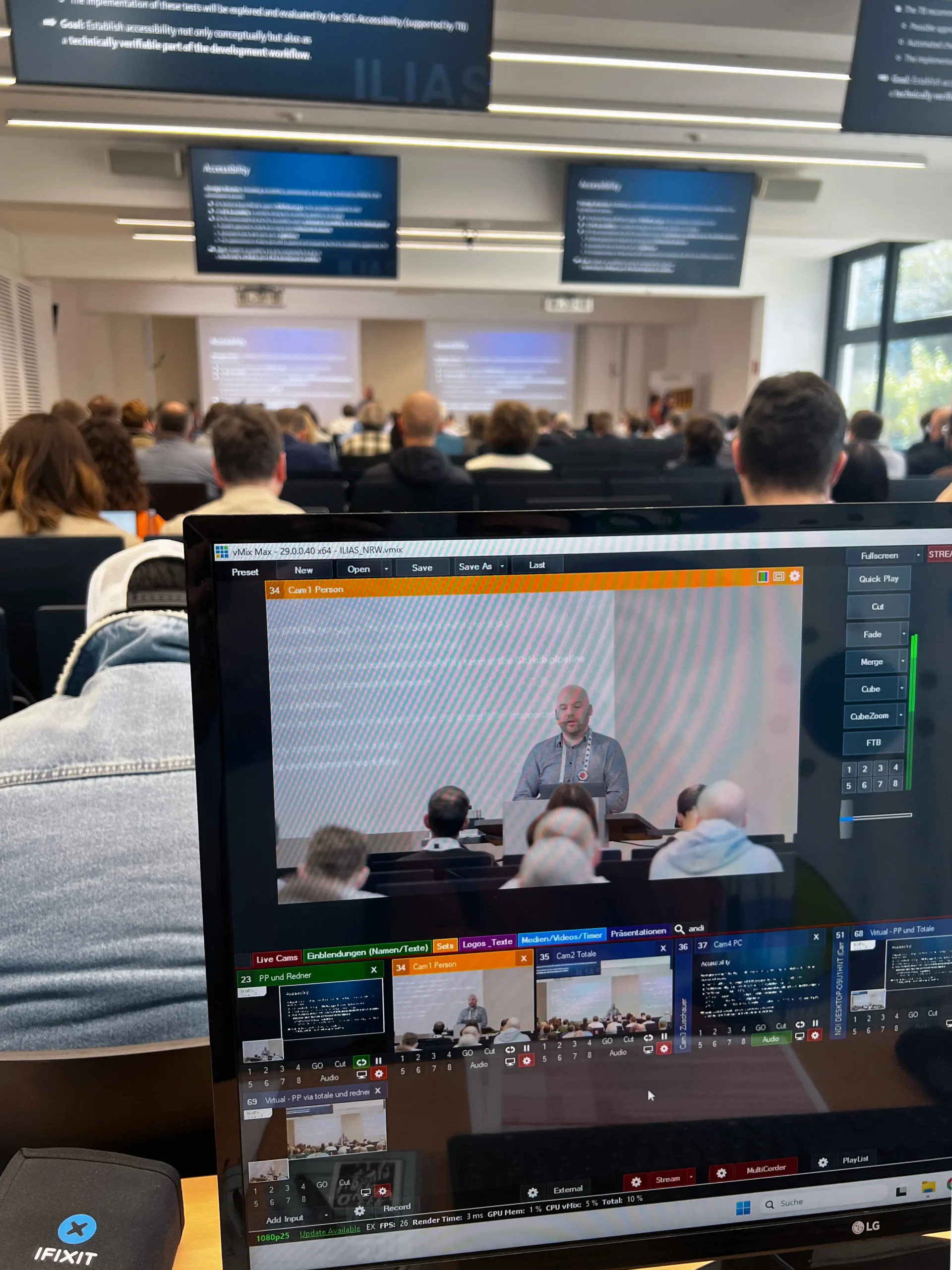 Foto: Blick von hinten auf einen Konferenzraum mit Publikum, im Vordergrund ein Bildschirm mit Videobearbeitungssoftware und mehreren Videovorschauen, auf denen ein Redner vor einer Leinwand mit Text zu sehen ist.