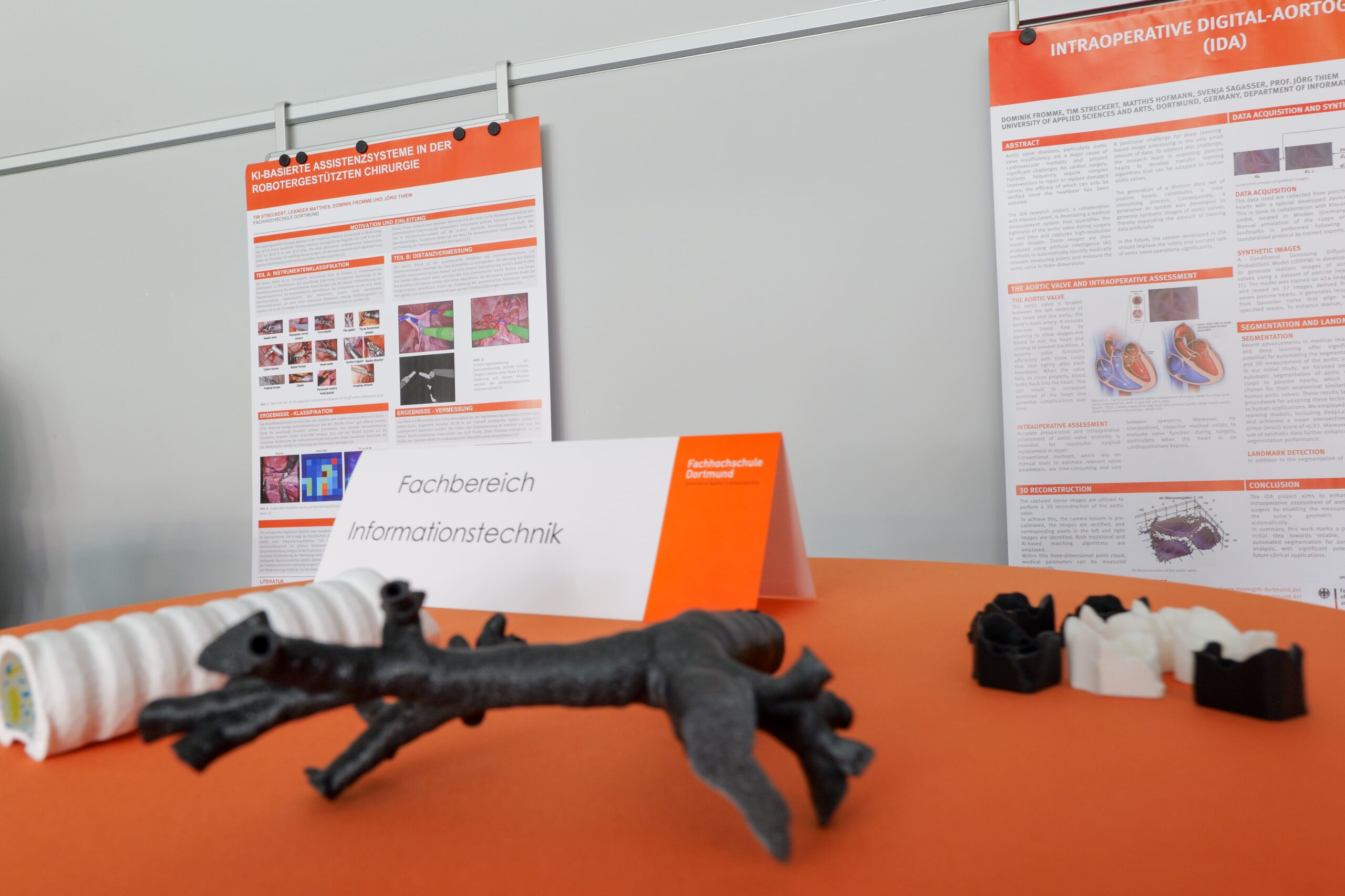 Foto: 3D-gedruckte Modelle von Blutgefäßen auf einem orangefarbenen Tisch vor Informationspostern und einem Schild mit der Aufschrift 'Fachbereich Informationstechnik'