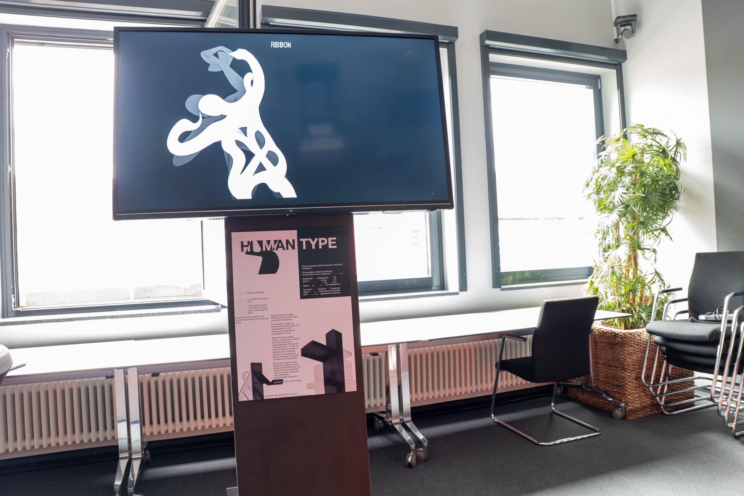 Foto: großer Bildschirm, der eine abstrakte weiße Figur auf dunklem Hintergrund zeigt, darunter ein Poster mit der Aufschrift "Human Type"