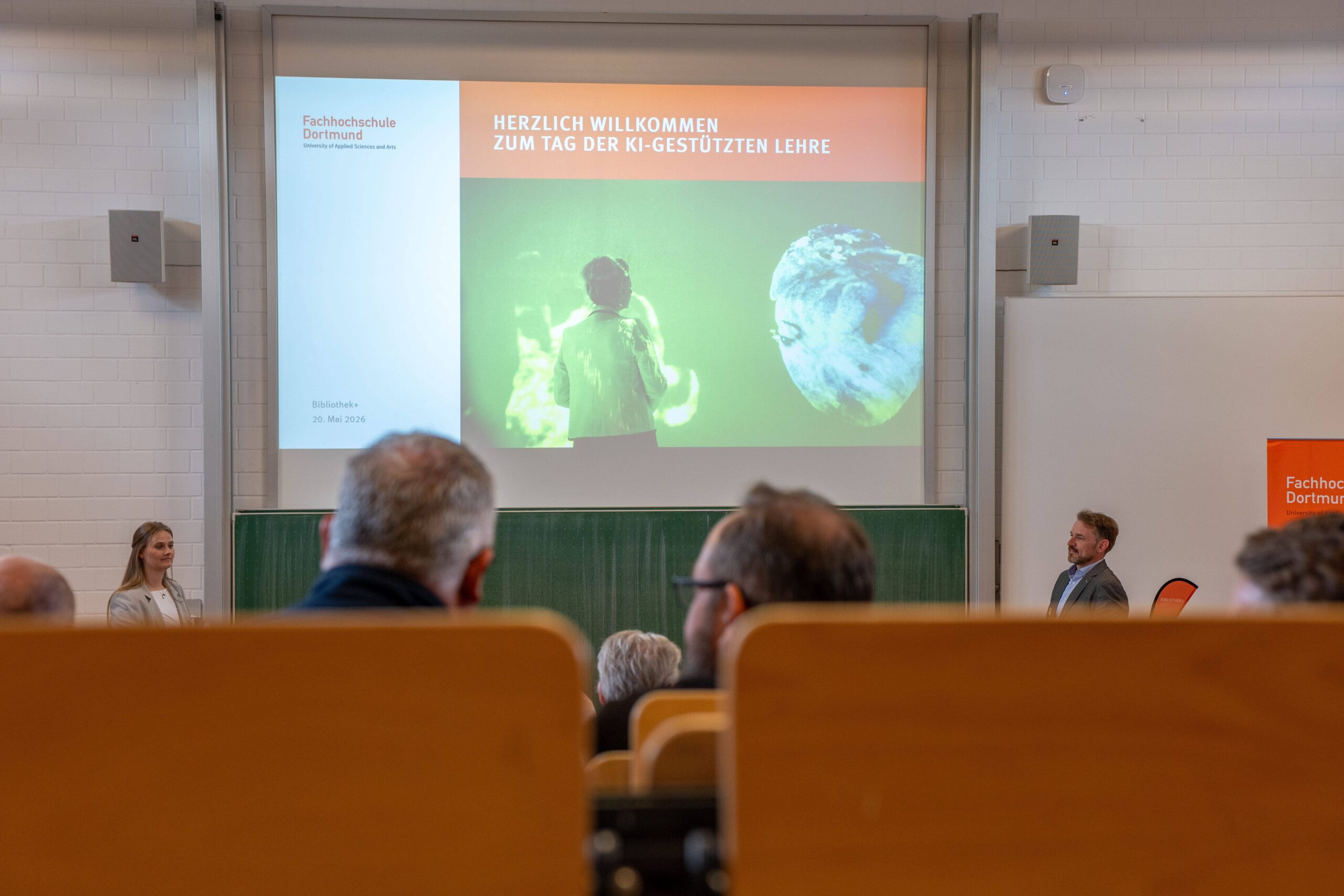 Foto: Blick aus den hinteren Reihen des Vortragsraums auf die Leinwand, dort ist zu lesen: "Herzlich Willkommen zum Tag der KI-gestützten Lehre"